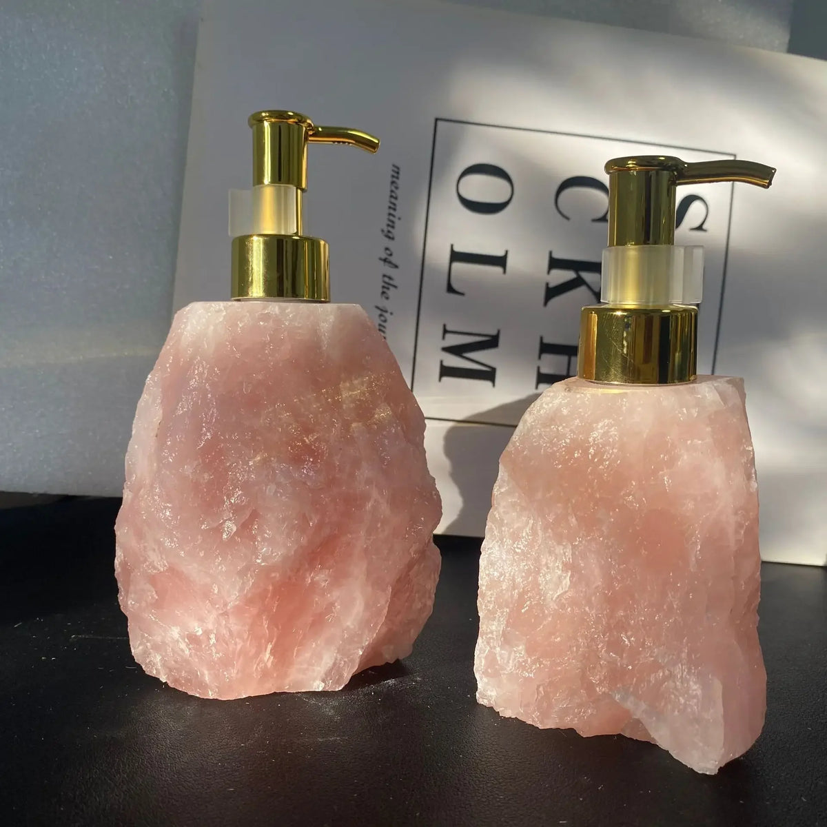 Two eprolo natural pink crystal stone liquid dispenser bottles with gold pumps.